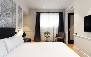 A hotel room with a large bed, a desk with a chair, a small table with a plant, and a TV on the wall. - Maldà Singular Hotel (Barcelona)