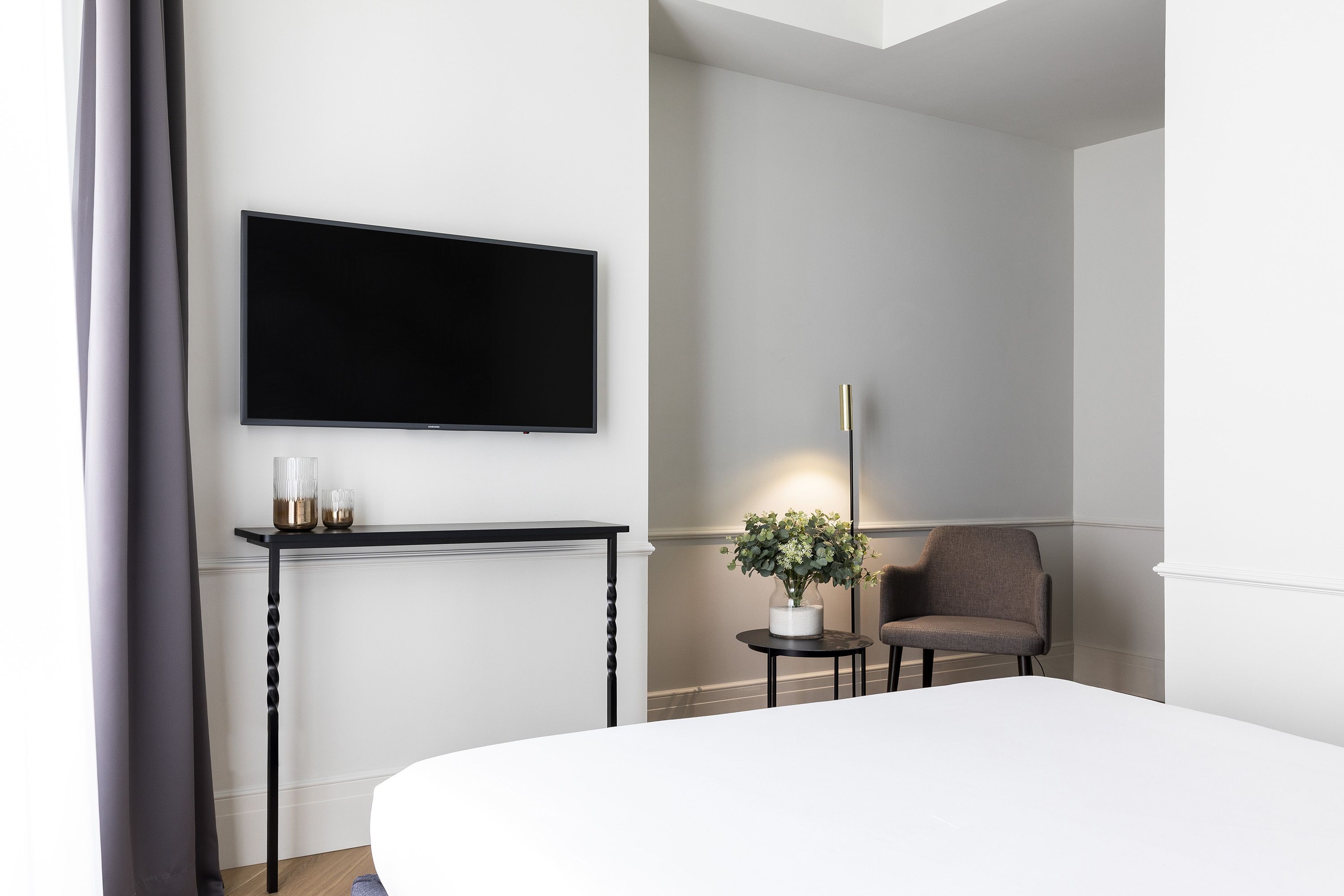 Een moderne hotelkamer met een groot bed, een flatscreen televisie aan de muur, een klein rond tafeltje met een bloevas en een stoel.