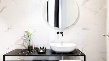 Un baño moderno con pared de mármol, espejo redondo, tocador negro, lavamanos blanco y un pequeño jarrón con flores.