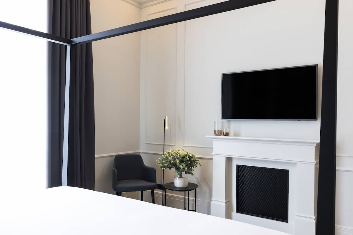 A modern hotel room with a large bed, a flat-screen TV mounted above a fireplace, a small table with a vase of flowers, and a dark armchair.