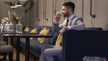A man seated on a dark blue sofa with yellow pillows, in a room with a wooden floor and a table with a glass of whiskey.
