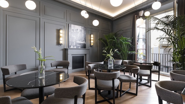A modern dining area with round tables and chairs, a fireplace, and large windows with plants. - Maldà Singular Hotel (Barcelona)