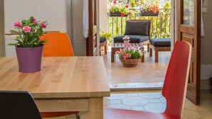 Une pièce avec une table en bois, un pot de fleurs violet et une chaise rouge. Il y a une fenêtre donnant sur un balcon agrémenté de plantes et d’une chaise.