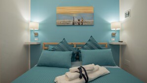 Un lit bien fait, avec une literie et des oreillers bleus, un tableau représentant une scène de plage accroché au mur, et deux lampes de chevet.
