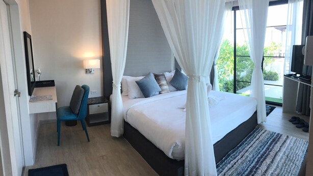 A bedroom with a bed, a bedside table, a chair, a TV, and a view of greenery outside. - La Playa Beach Front Pool Villa Cha-am (Cha-am)