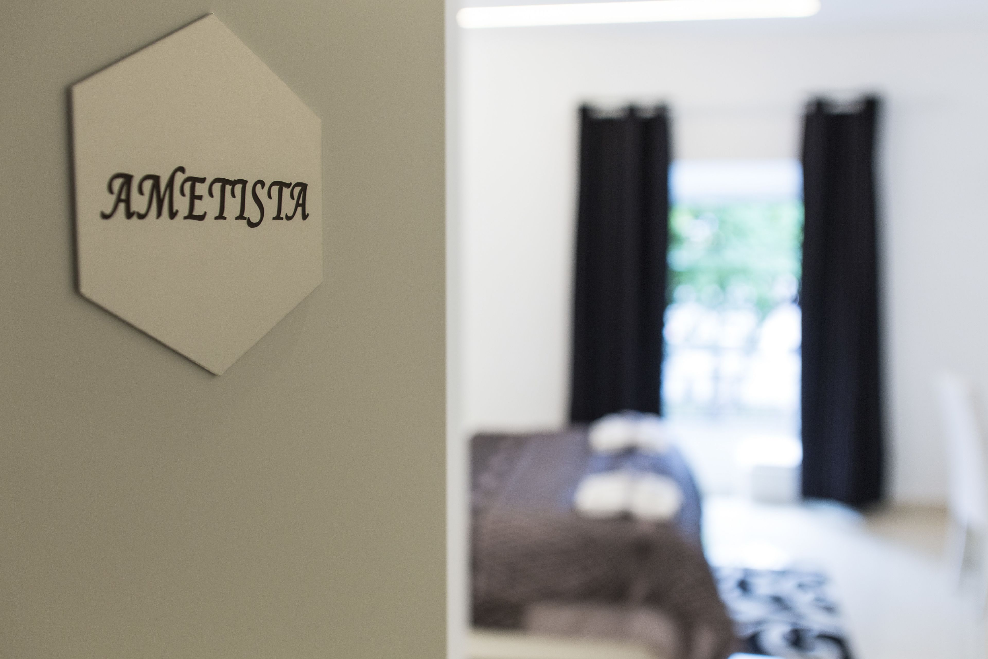 Un panneau hexagonal sur lequel est inscrit le mot « AMETISTA », fixé à un mur, et une pièce floue avec un lit et des rideaux.