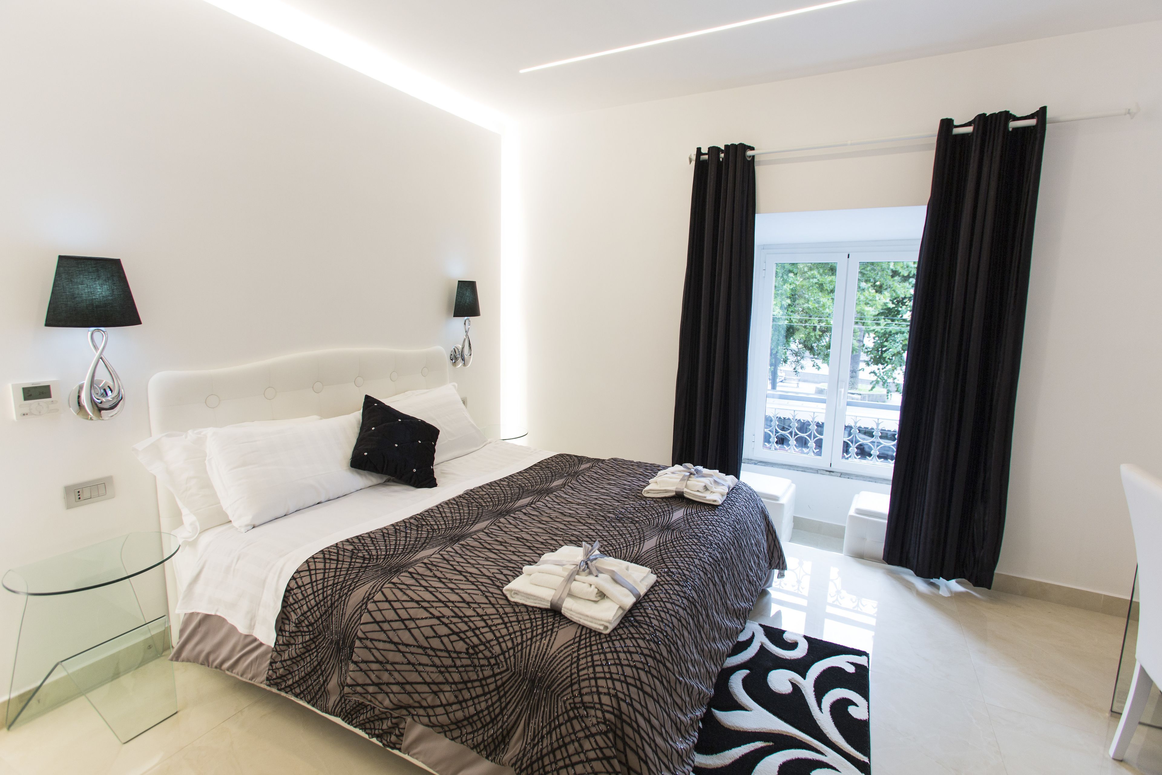 Una habitación de hotel moderna con una cama grande, dos lámparas de noche, una ventana con cortinas y una mesita de vidrio.