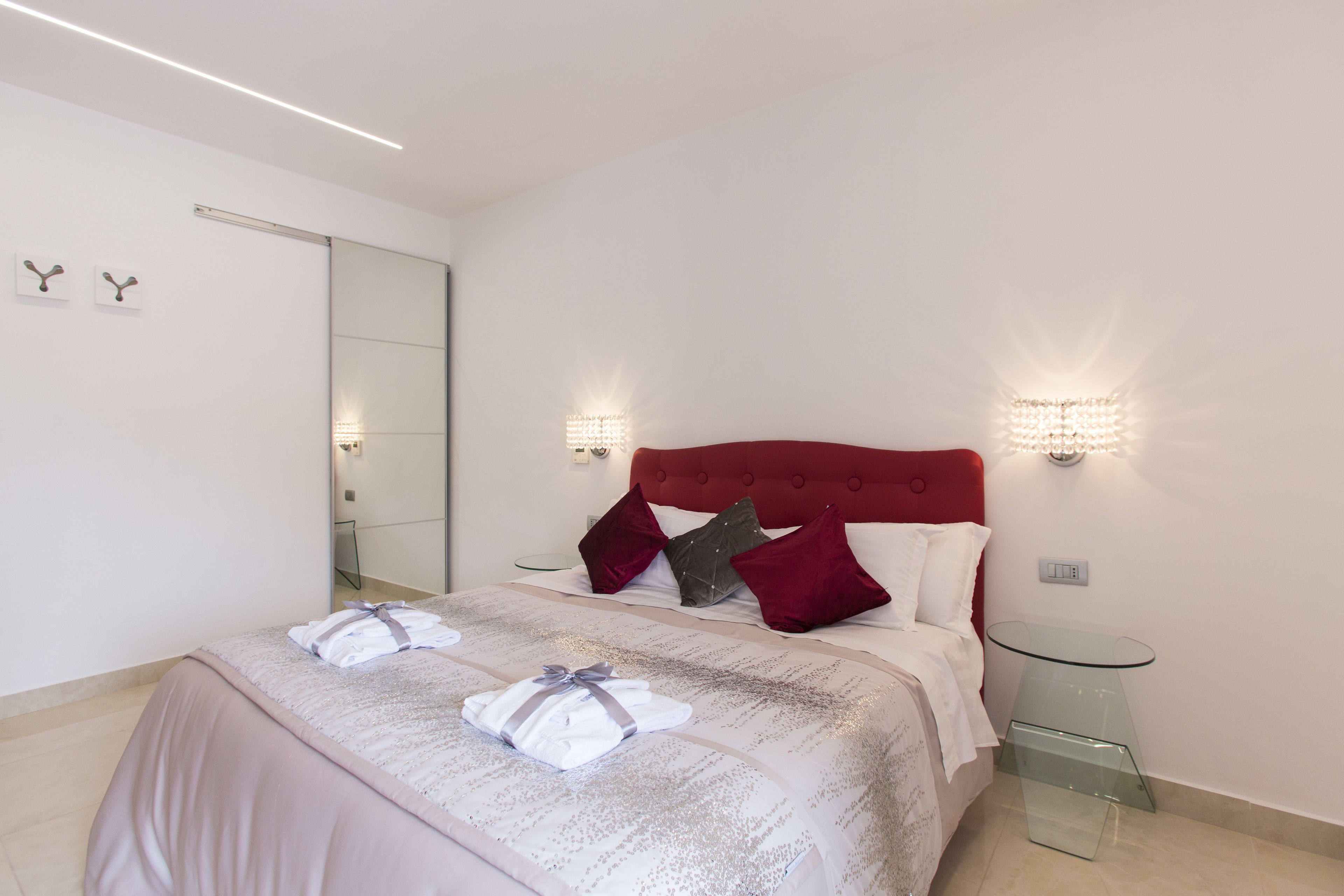 A hotel room with a large bed, a glass side table, a red headboard, and two wall-mounted light fixtures.