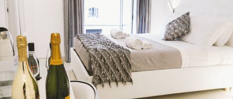 A modern hotel room with a bed, a bedside table with two bottles of champagne, and a view of the city through the window.