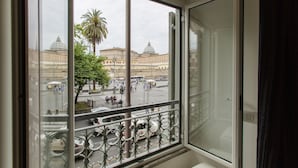 A room with a large window offering a view of a city square with historic buildings and palm trees. - San Pietro Grand Suite (Rome)
