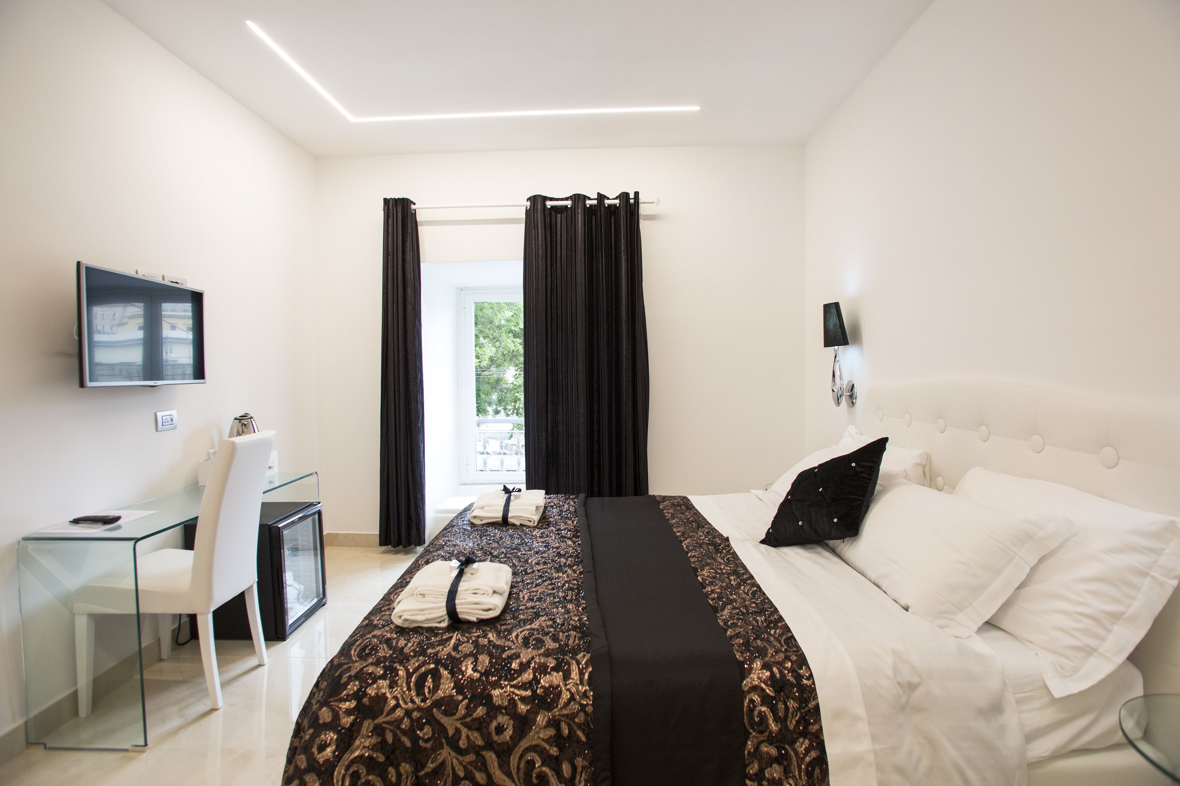 Habitació d'hotel amb un llit, una taula de treball amb cadira, un televisor i una finestra amb cortines.