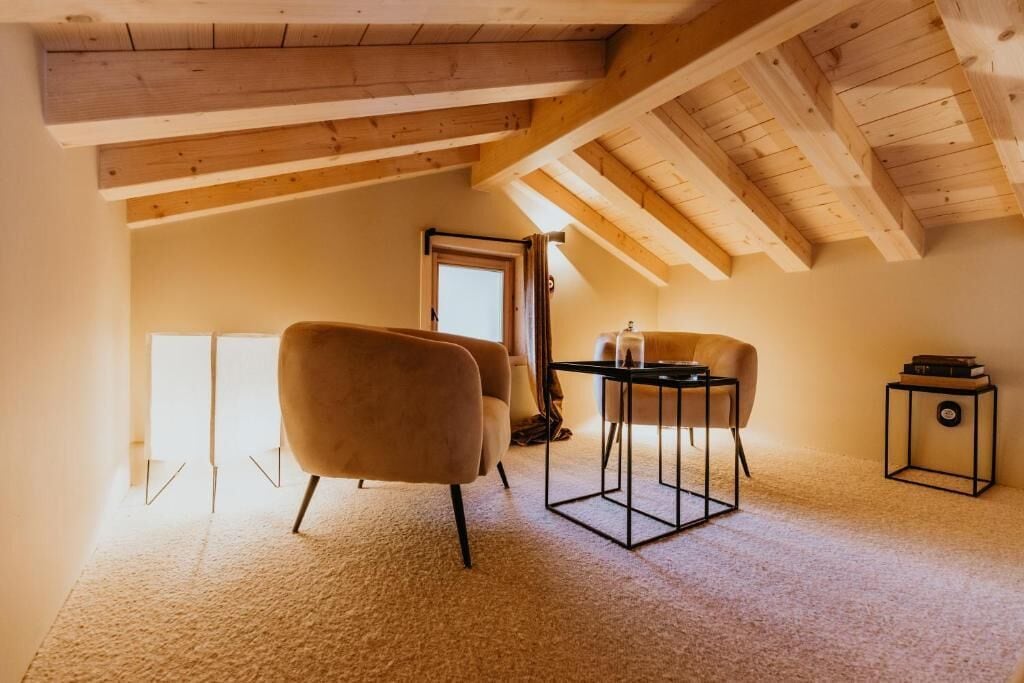 Una habitación con techo alto, vigas de madera, piso alfombrado, una mesa con sillas y una puerta que da al exterior.