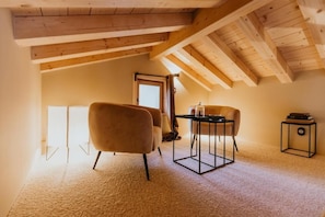 A room with a high ceiling, wooden beams, a carpeted floor, a table with chairs, and a door leading outside.