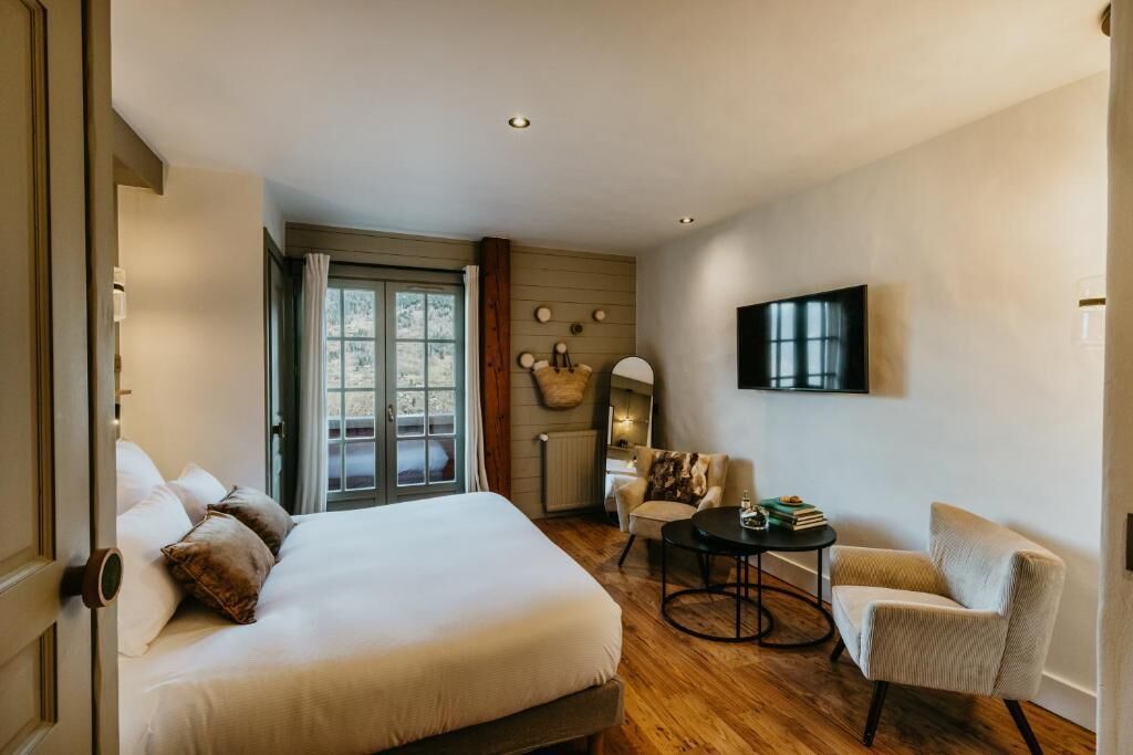 Una habitación de hotel con una cama grande, una mesa redonda, un sofá, un televisor y un ventanal con cortinas.