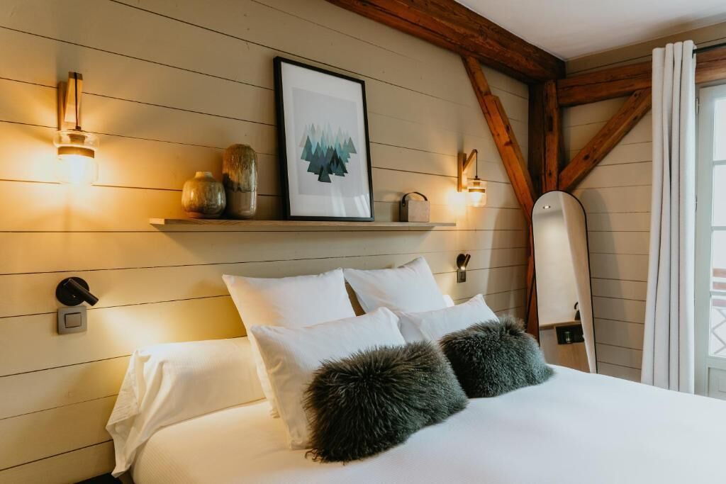 Een slaapkamer met een bed, kussens, wandlampen, een ingelijst schilderij en een houten plafond.