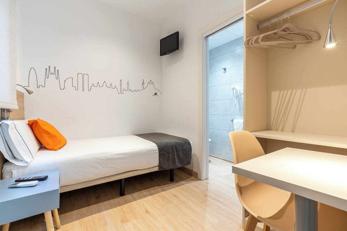 A modern hotel room with a bed, a desk, a chair, a TV, and a city skyline wall art.