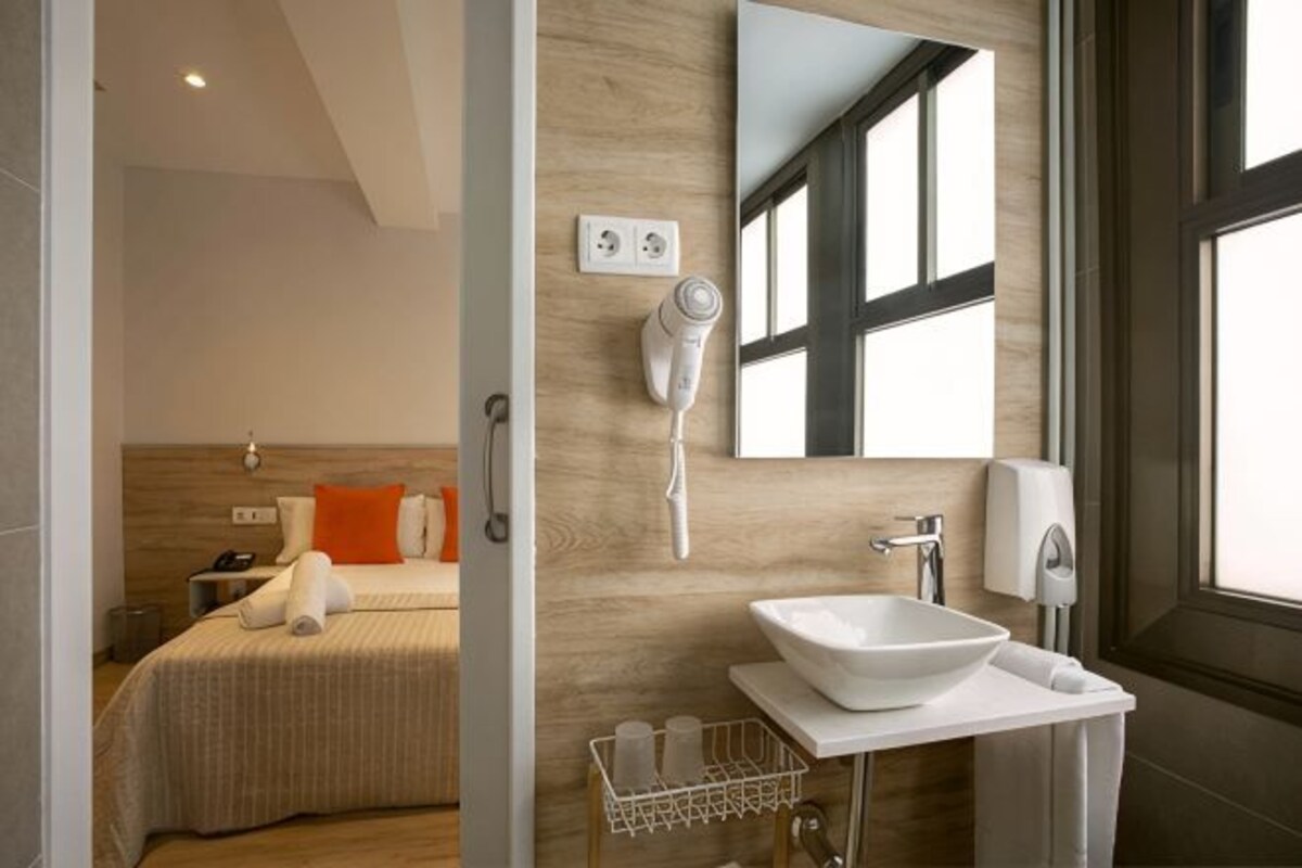 A modern hotel room with a bed, a bathroom with a sink and mirror, and a hairdryer on the wall.