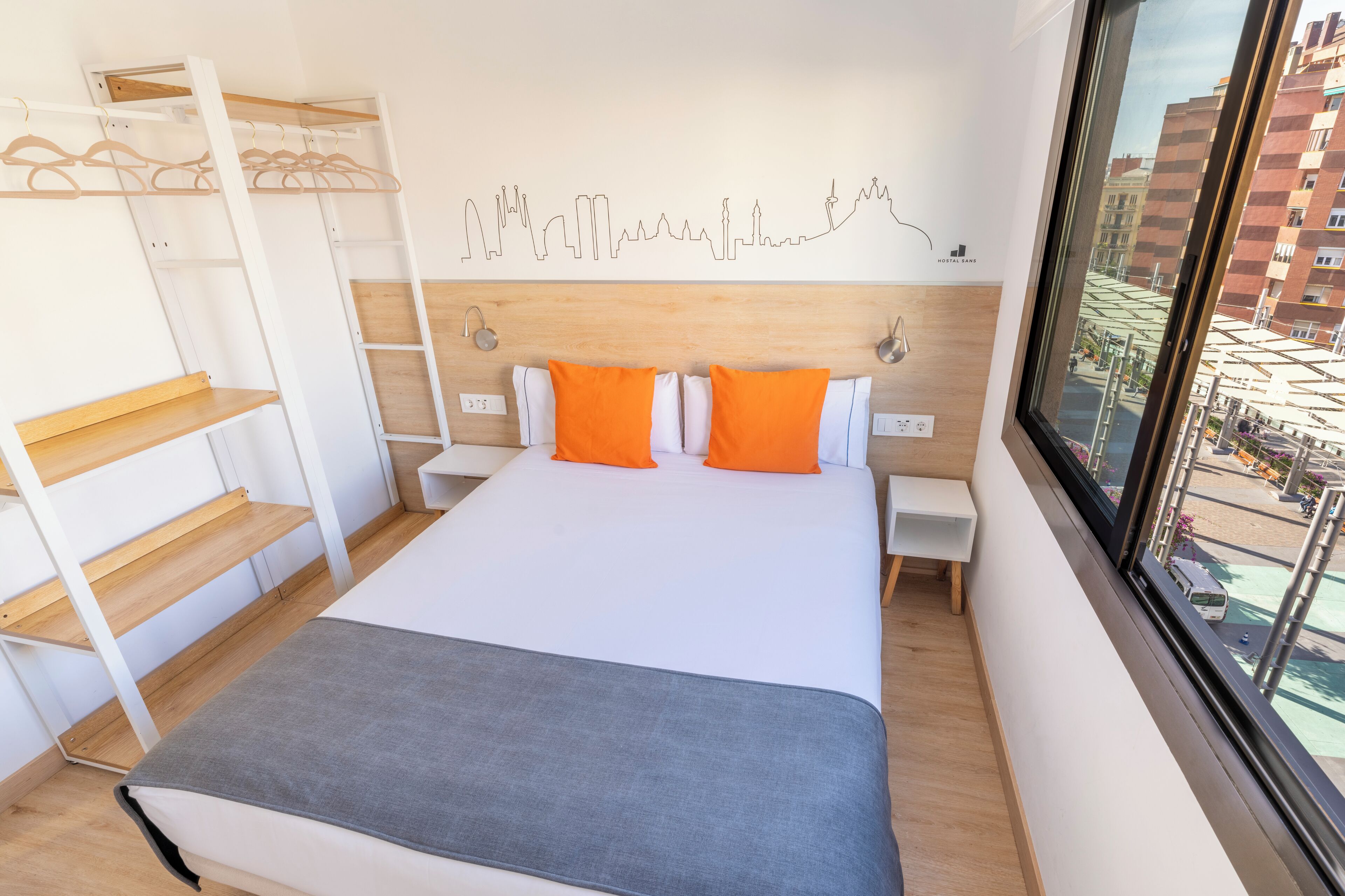 A modern hotel room with a large bed, a wooden headboard, a city skyline view, and a built-in wardrobe with shelves.