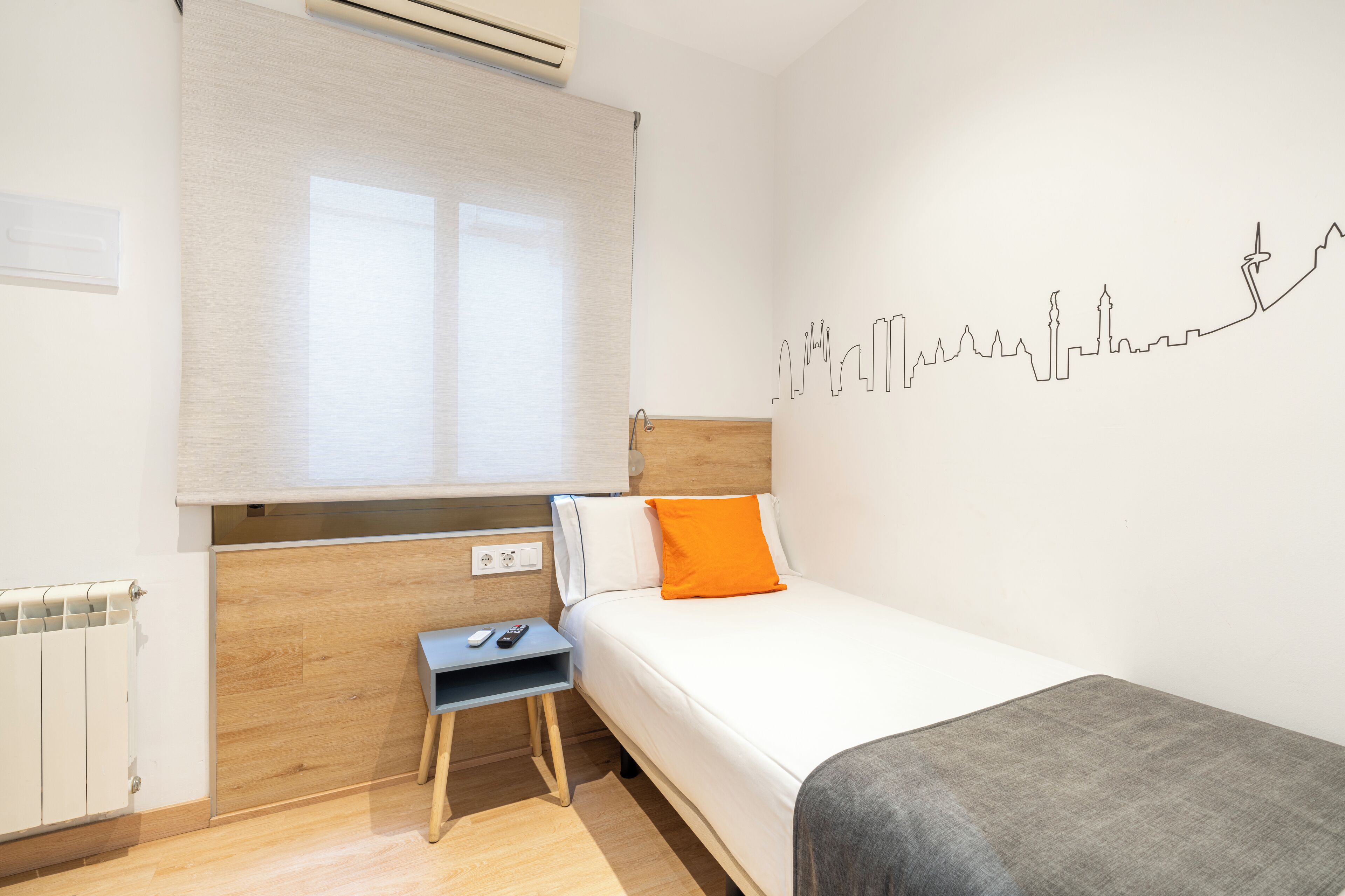A modern hotel room with a bed, a small desk, and a wall with a city skyline mural.