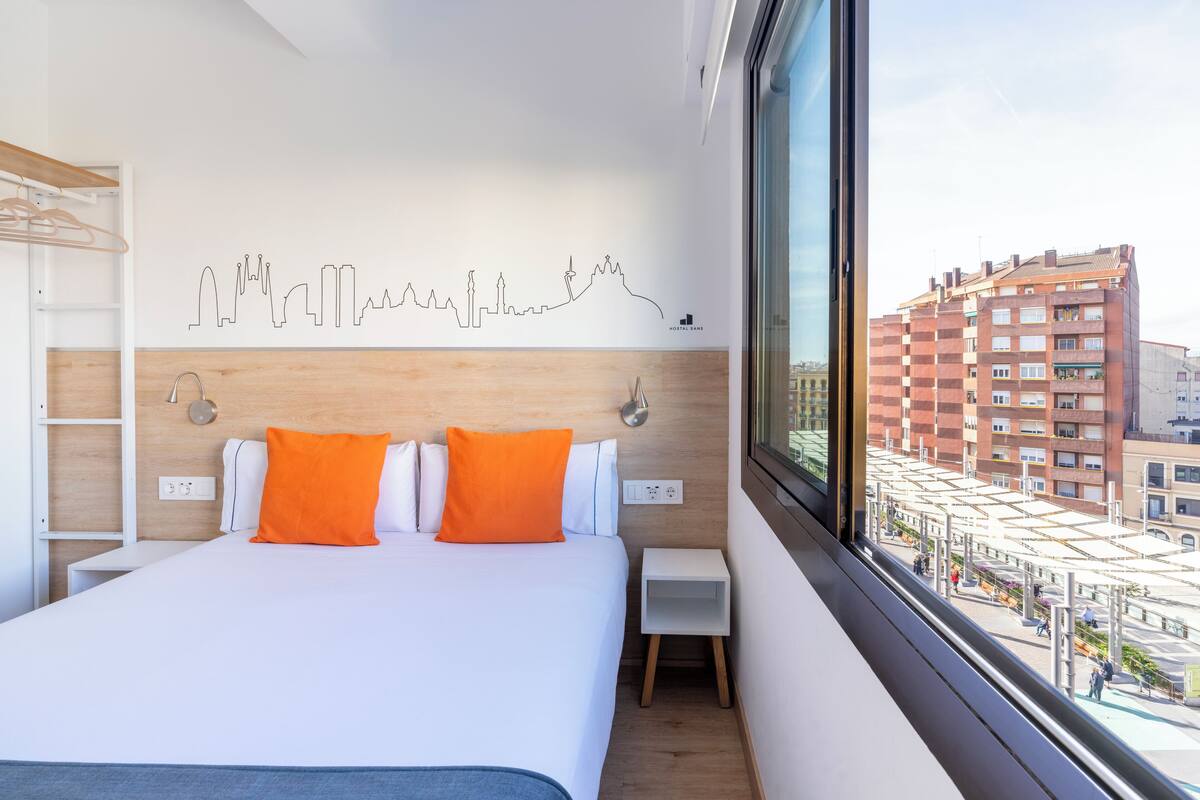 A modern hotel room with a city skyline wall art, a bed with white and orange bedding, a small bedside table, and a window with a view of the cityscape.