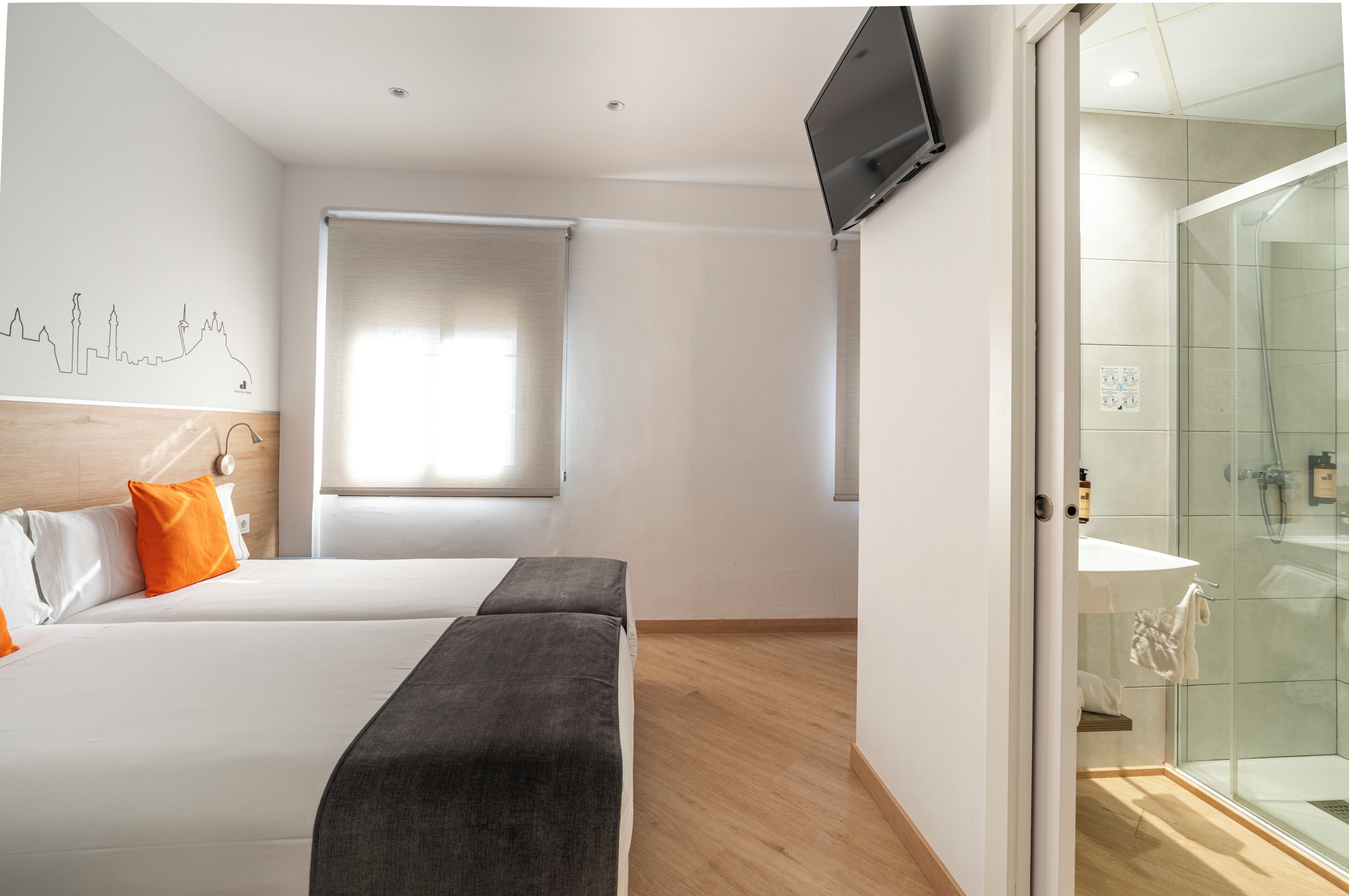 Habitación de hotel con cama, televisor montado en la pared, ventana con persianas y zona de ducha de vidrio.