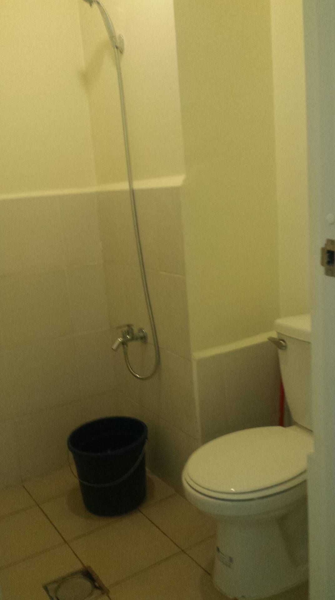 Une petite salle de bain avec des toilettes, une douche et un seau noir posé sur le sol.