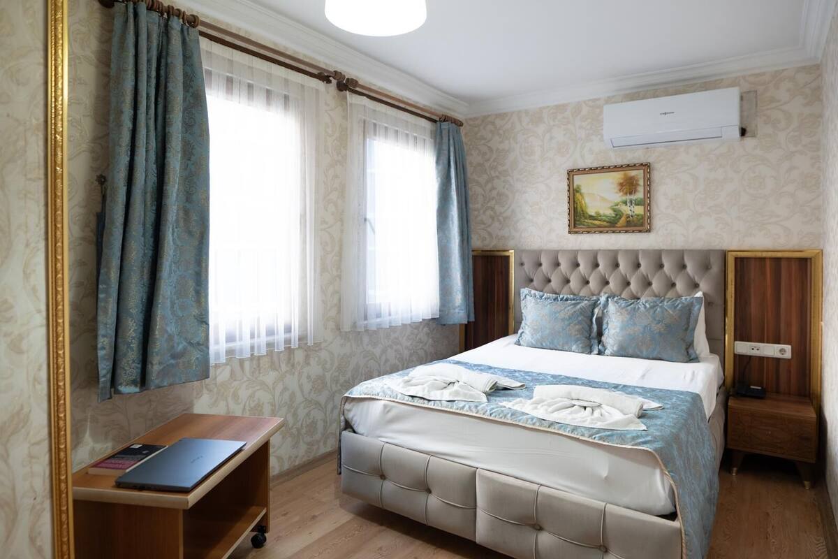 A bedroom with a tufted headboard, a bed with blue and white bedding, a wooden nightstand, a desk with a laptop, and a painting on the wall.