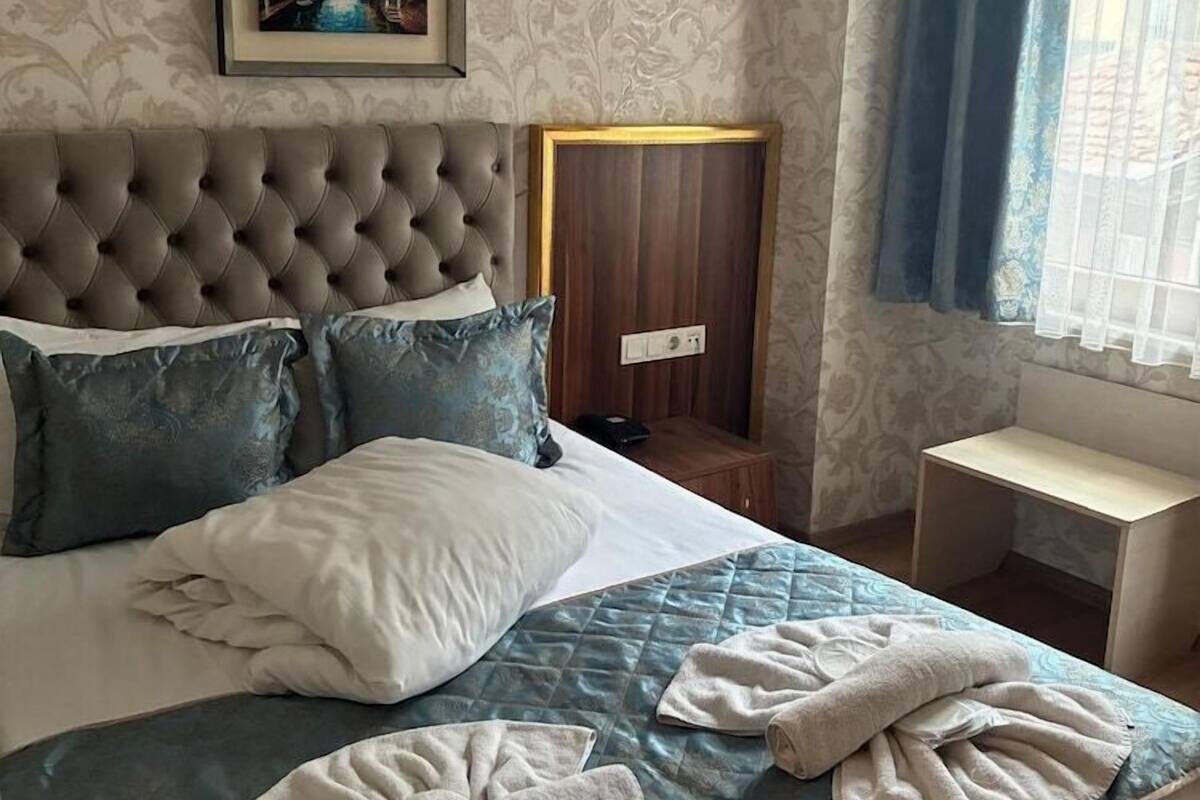 A bedroom with a tufted headboard, a bed with blue and white bedding, a framed picture on the wall, a wooden nightstand, and a window with blue curtains.