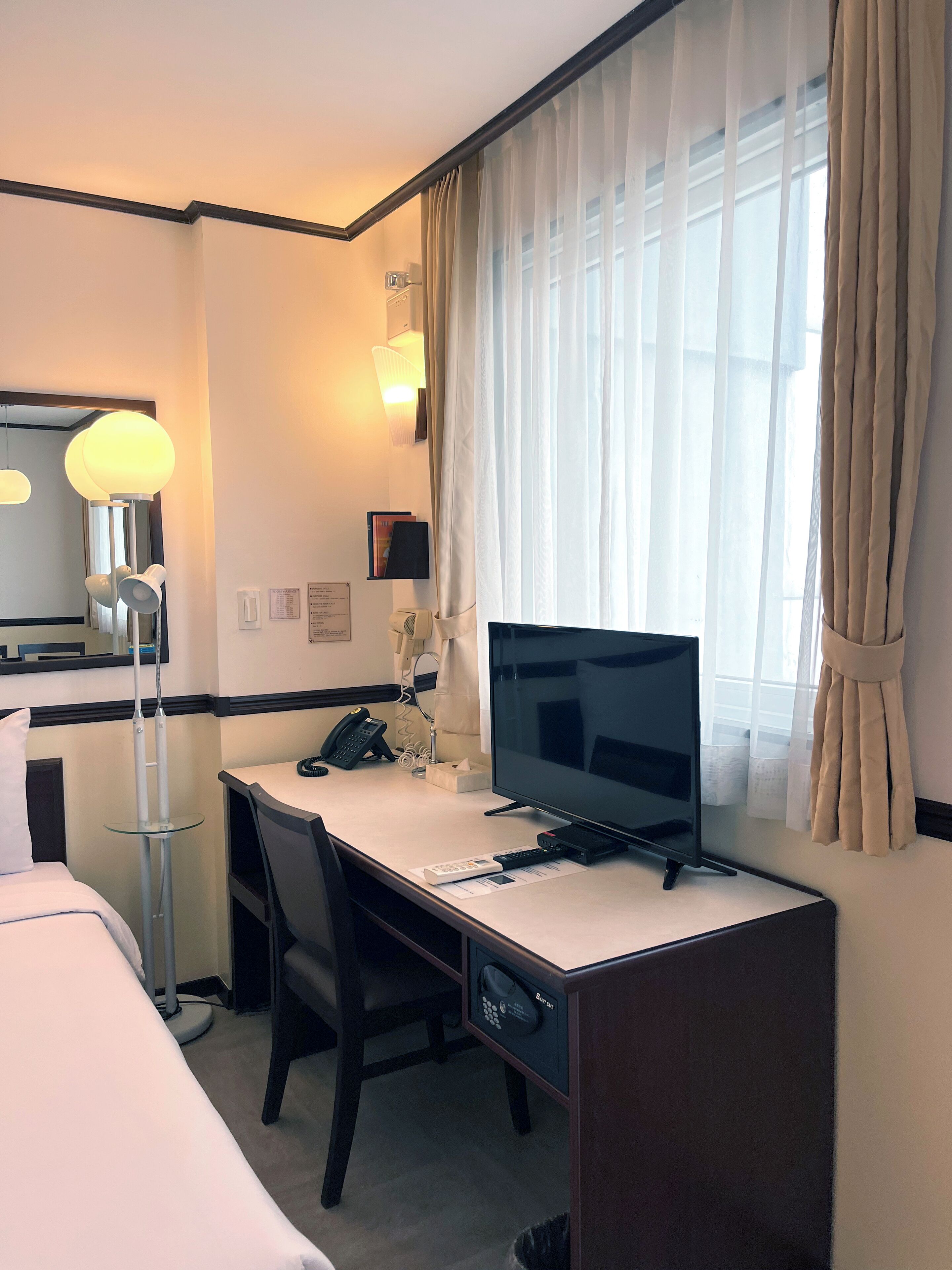 Habitación de hotel con cama, escritorio, computadora, silla, lámpara, teléfono y ventana con cortinas.