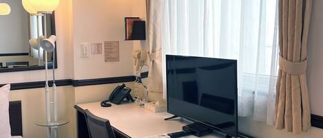 A hotel room with a bed, a desk with a computer, a chair, a lamp, a phone, and a window with curtains.