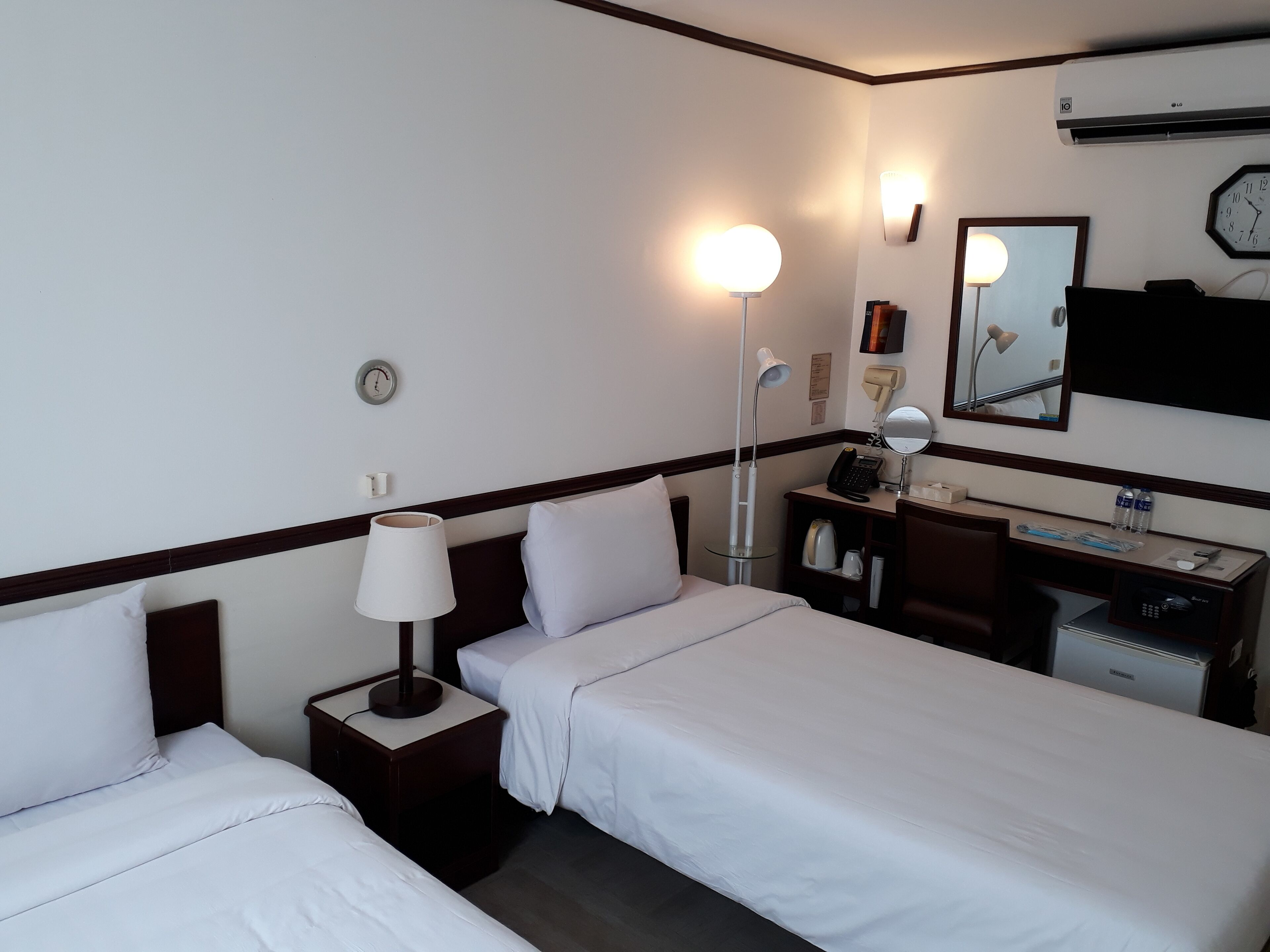 A hotel room with two single beds, a desk with a computer, a lamp, a clock, and a mirror.