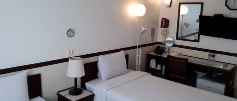 A hotel room with two single beds, a desk with a computer, a lamp, a clock, and a mirror.
