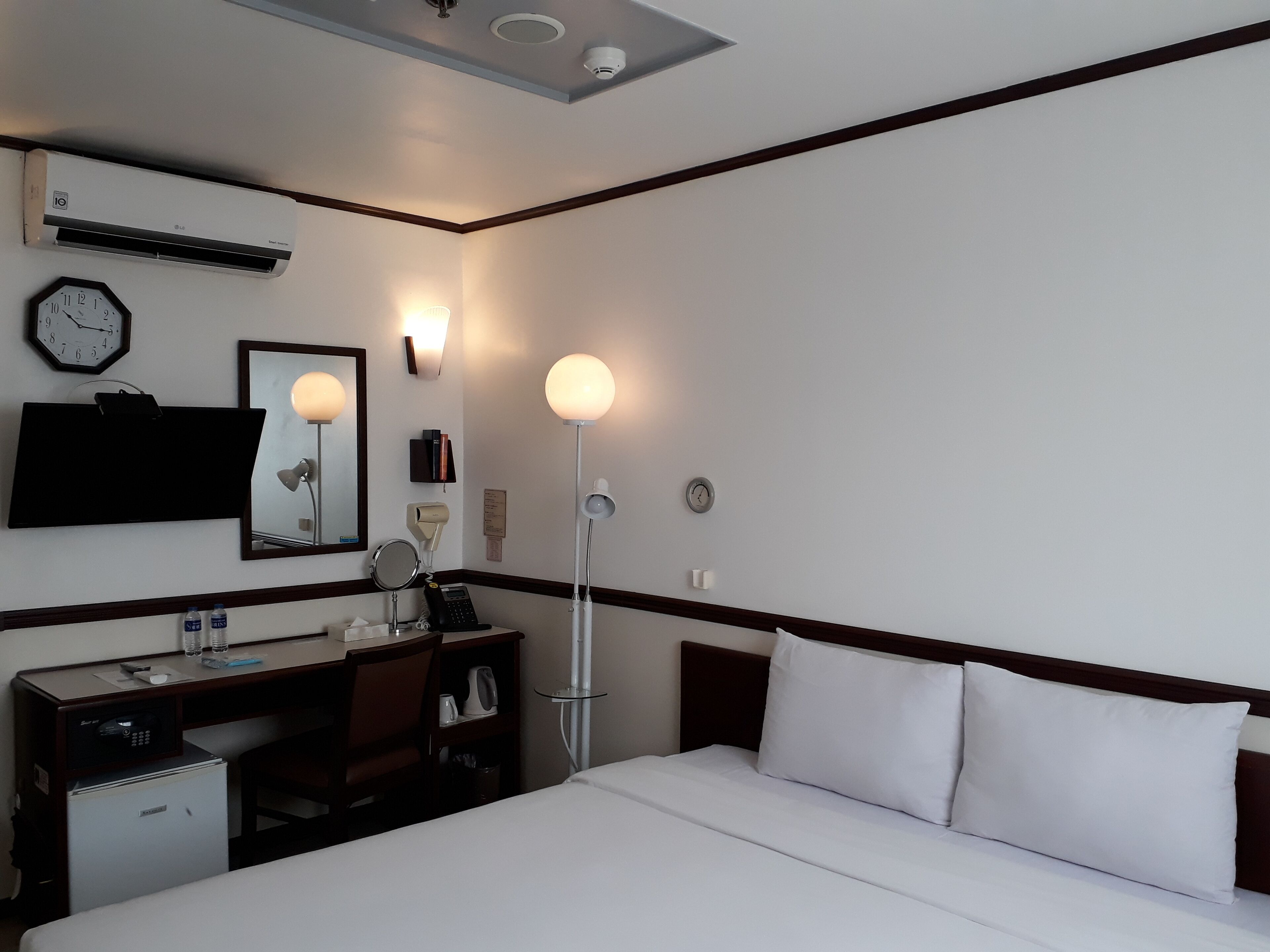 A hotel room with a bed, desk, and a mirror. There is a clock on the wall and a ceiling light fixture.