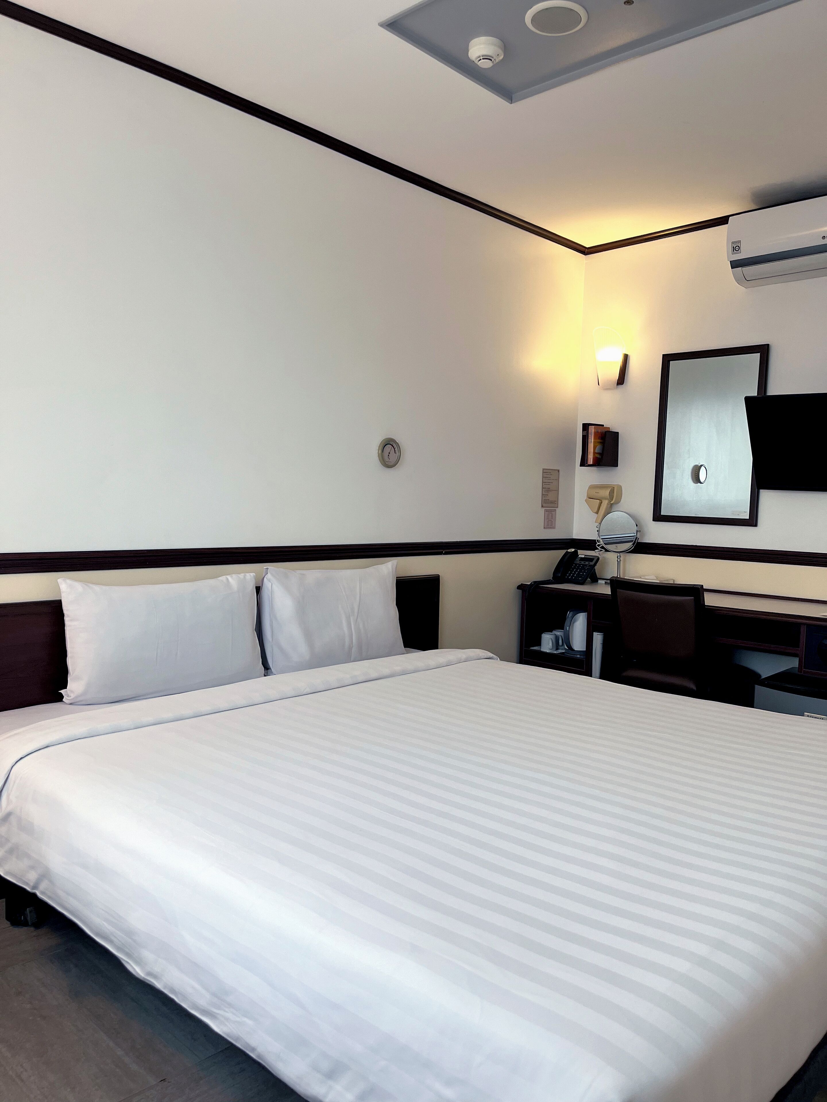 Una habitación de hotel con una cama grande, un escritorio con una silla, una lámpara de escritorio, un teléfono, un espejo y un equipo de aire acondicionado en la pared.