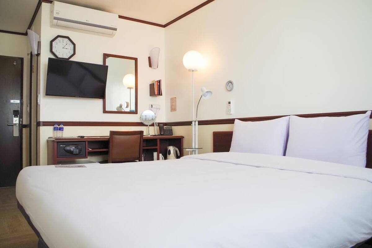 A hotel room with a large bed, a desk with a computer, a chair, a desk lamp, a clock, and a wall-mounted TV.