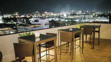 Una terraza en la azotea con mobiliario de mimbre, una mesa con menú y vistas a la ciudad por la noche.