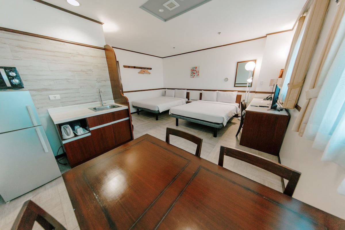 A compact hotel room with a wooden dining table, two single beds, a small kitchenette, and a mirror.