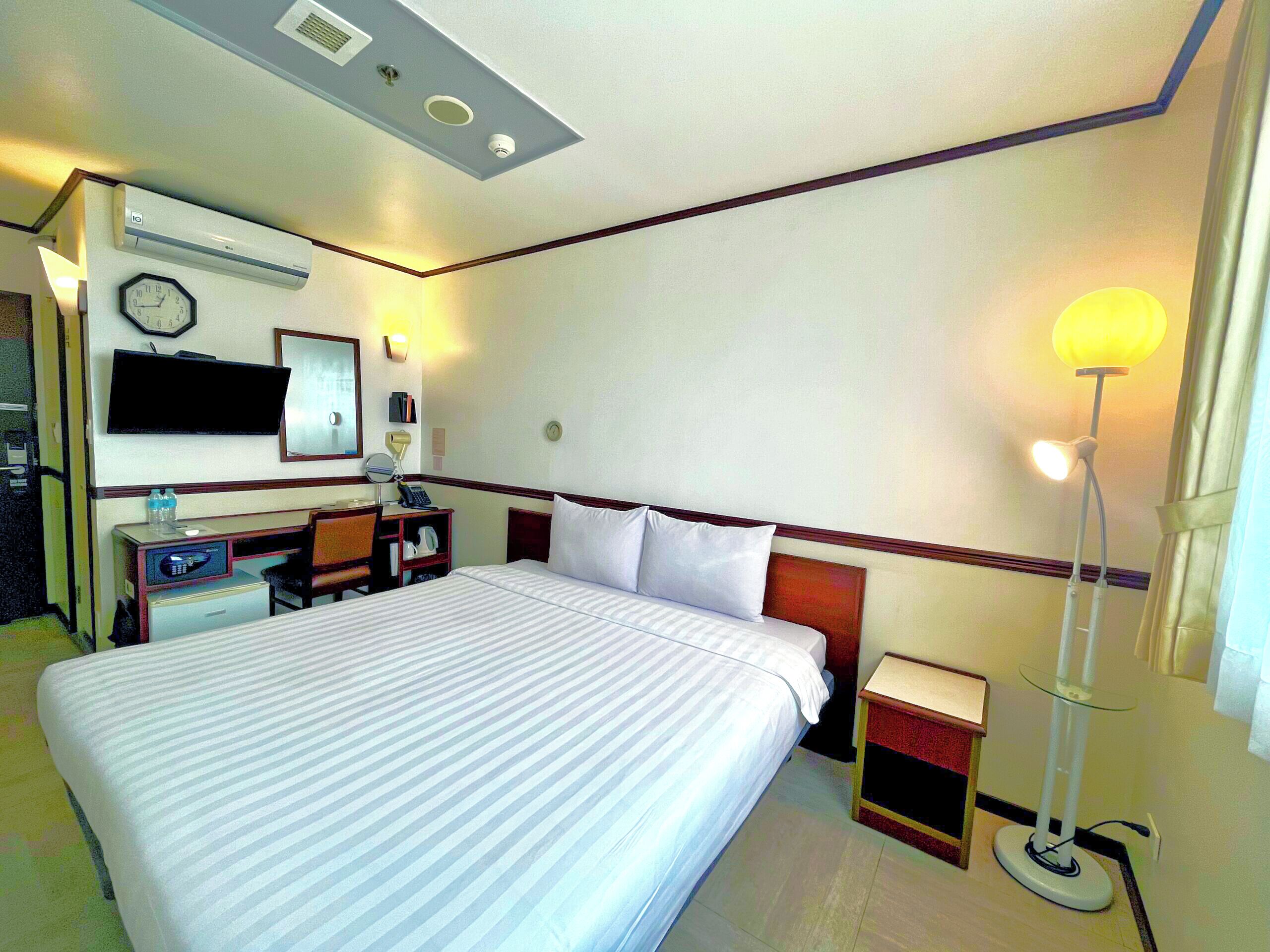 A hotel room with a large bed, a desk with a computer, a chair, a bedside table, a lamp, and a clock on the wall.