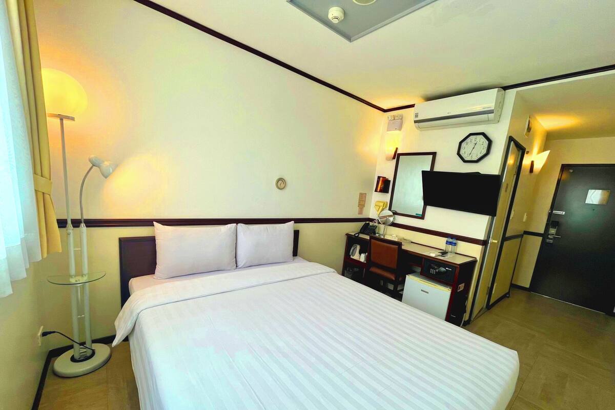 A hotel room with a bed, a desk with a chair, a lamp, a clock, and an air conditioning unit.