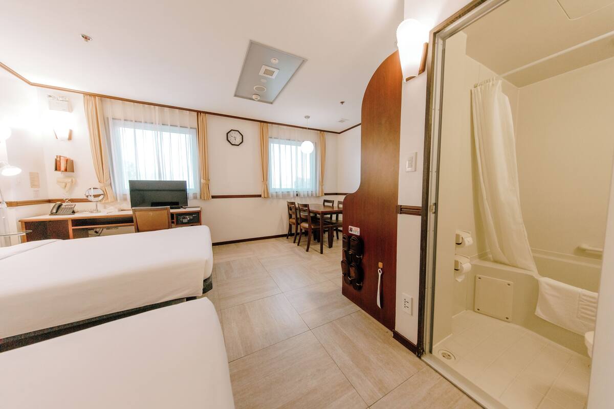 A hotel room with a bed, desk, chair, TV, and a shower area.
