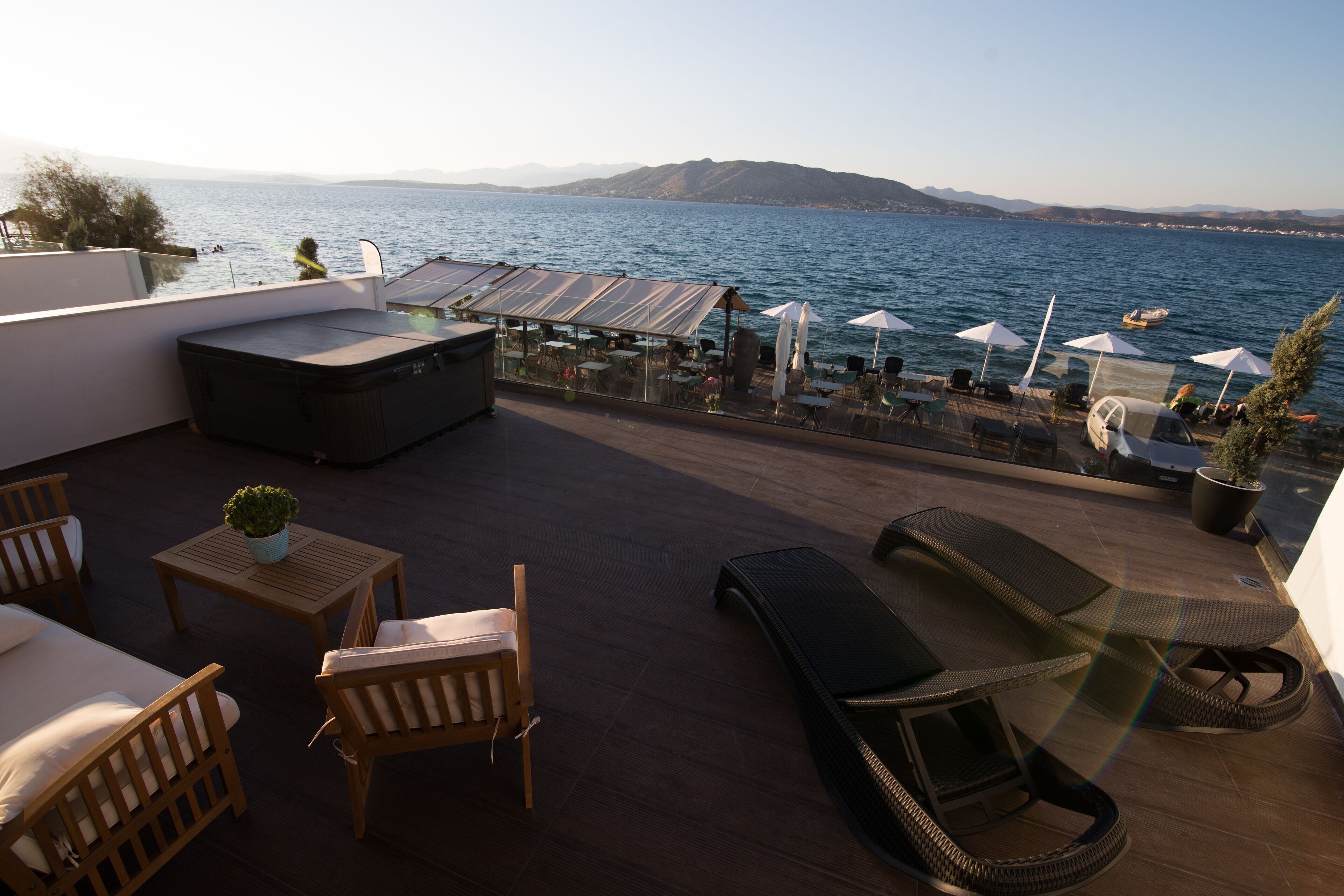 Terraza en la azotea con mobiliario de madera, una mesa grande y sombrillas, con vistas a un cuerpo de agua y montañas al fondo.