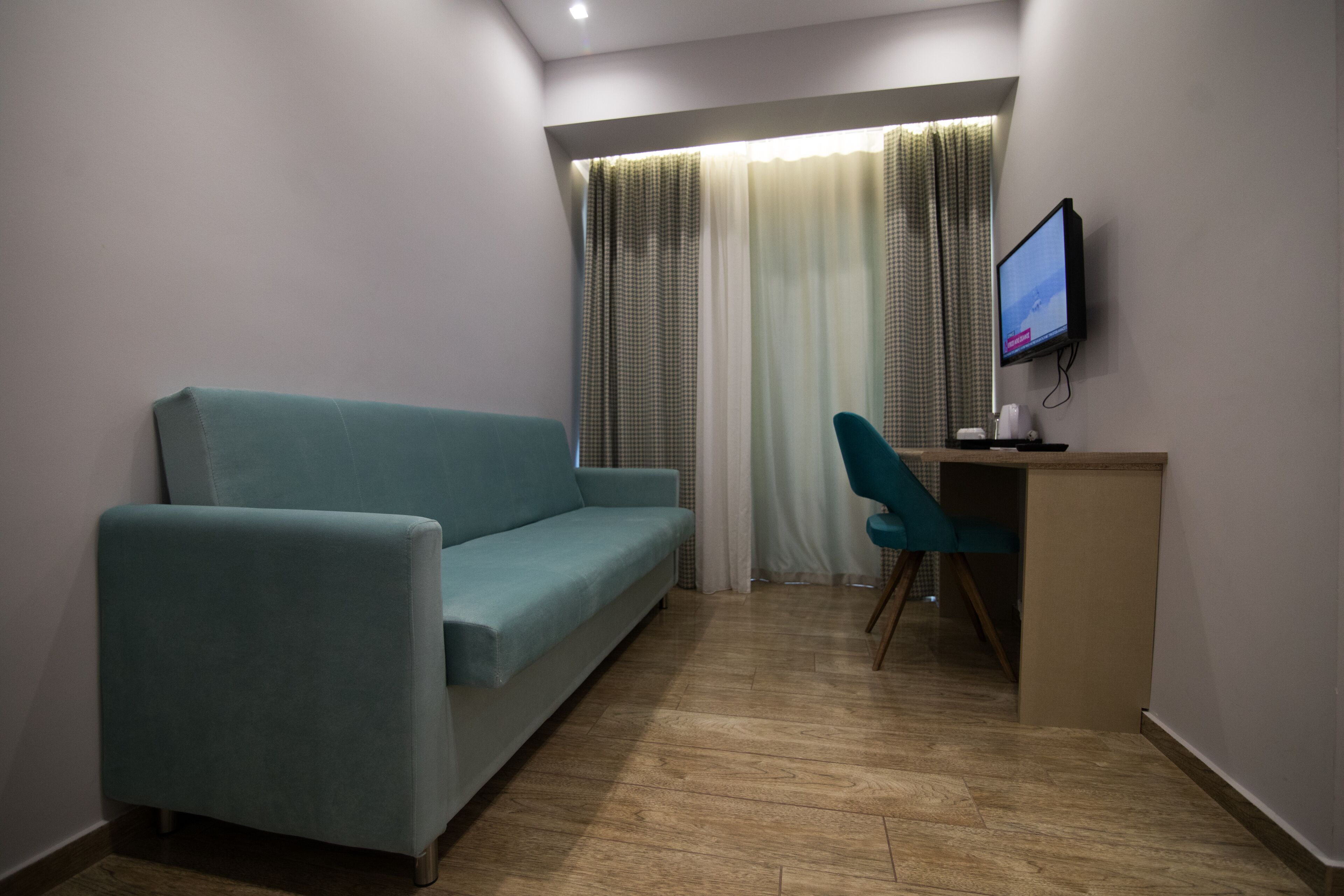 A room with a light blue sofa, a teal chair, a small desk with a monitor, and a window with curtains.