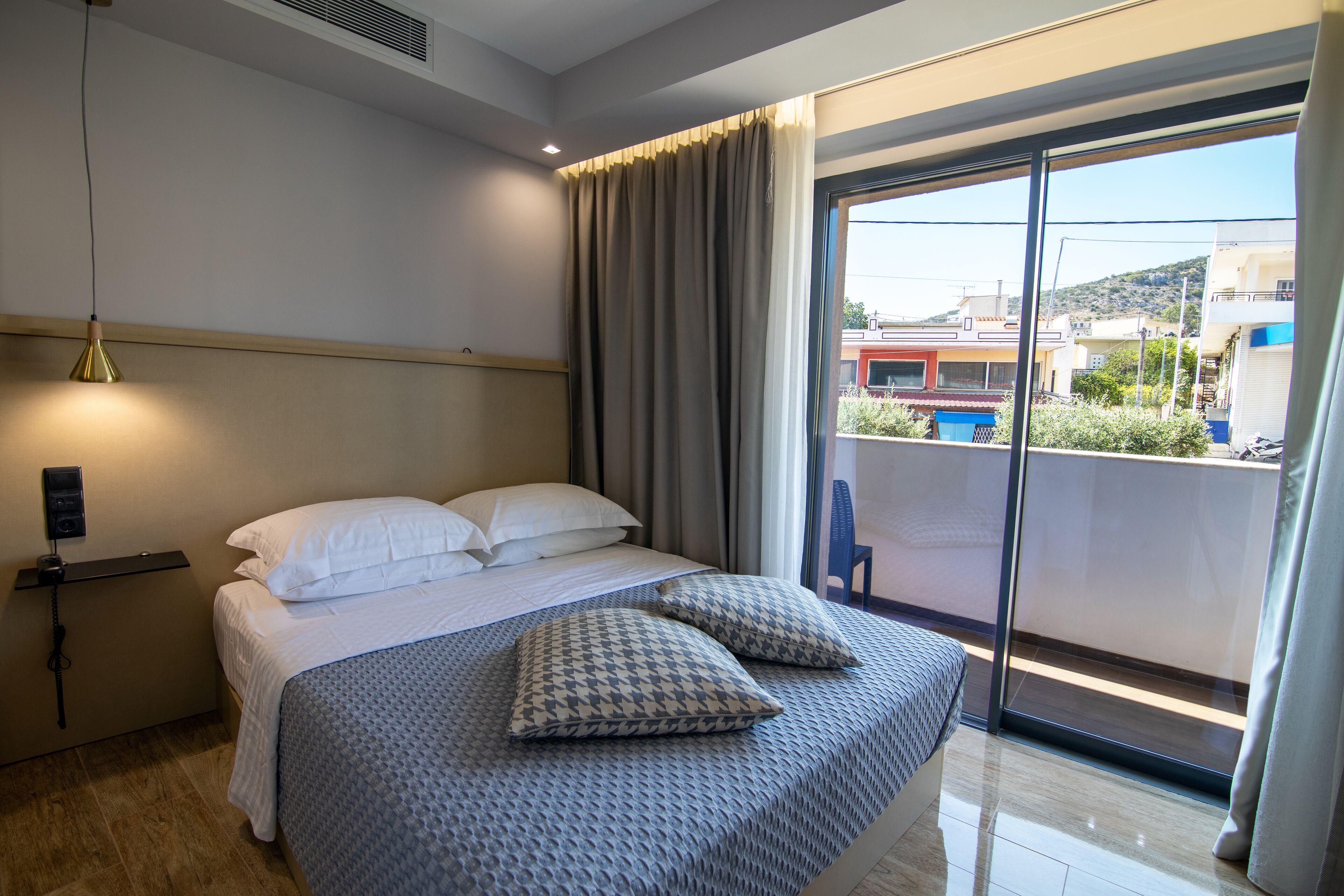 Habitación de hotel con cama, mesita de noche con lámpara, ventana con vista a edificios y balcón con una silla.