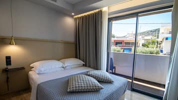 Habitación de hotel con cama, mesita de noche con lámpara, ventana con vista a edificios y balcón con una silla.