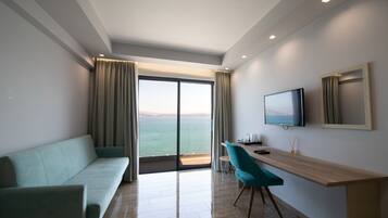 Una habitación de hotel moderna con sofá, un escritorio de madera, un televisor de pantalla plana y un amplio ventanal corredero que da a un balcón con vistas al mar.