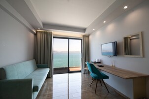 Una habitación de hotel moderna con sofá, un escritorio de madera, un televisor de pantalla plana y un amplio ventanal corredero que da a un balcón con vistas al mar.