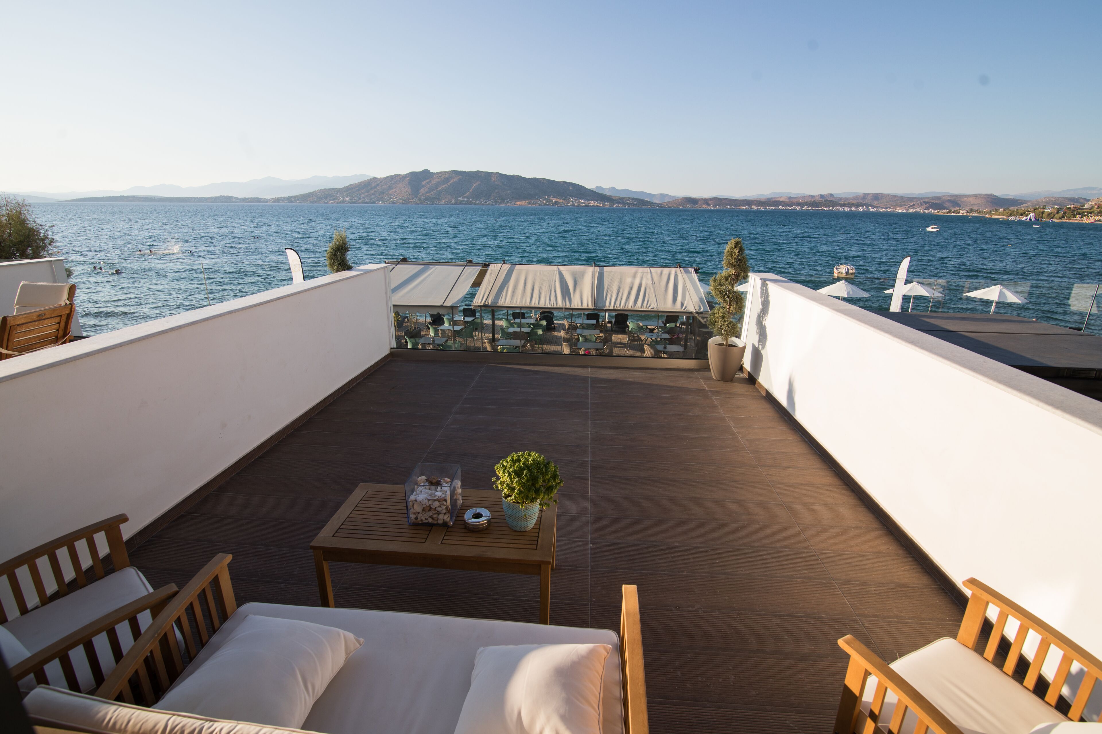 Una terraza con muebles de madera, una mesa con una planta y vistas al mar y montañas distantes.