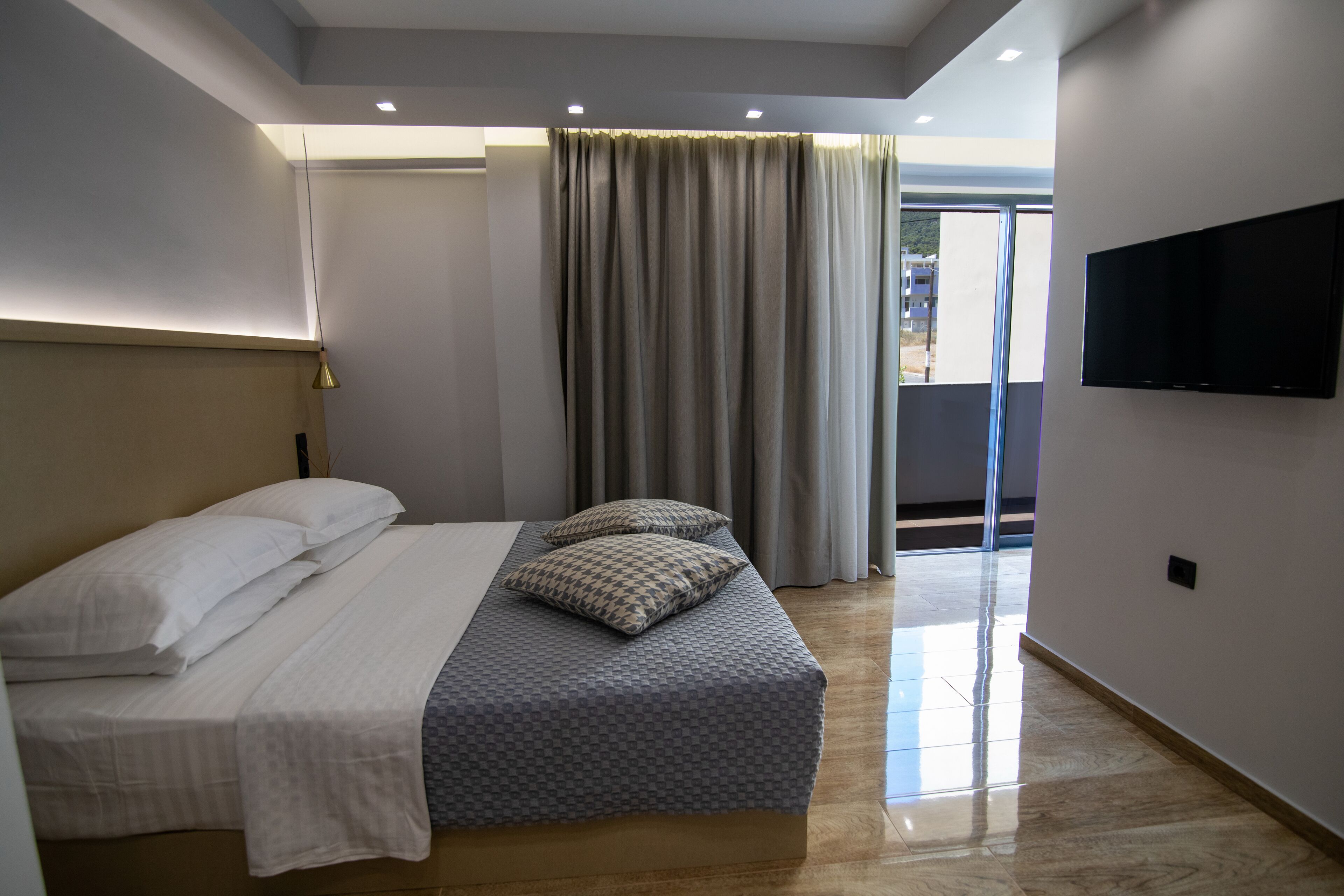 A modern hotel room with a large bed, a flat-screen TV mounted on the wall, and a balcony with curtains.