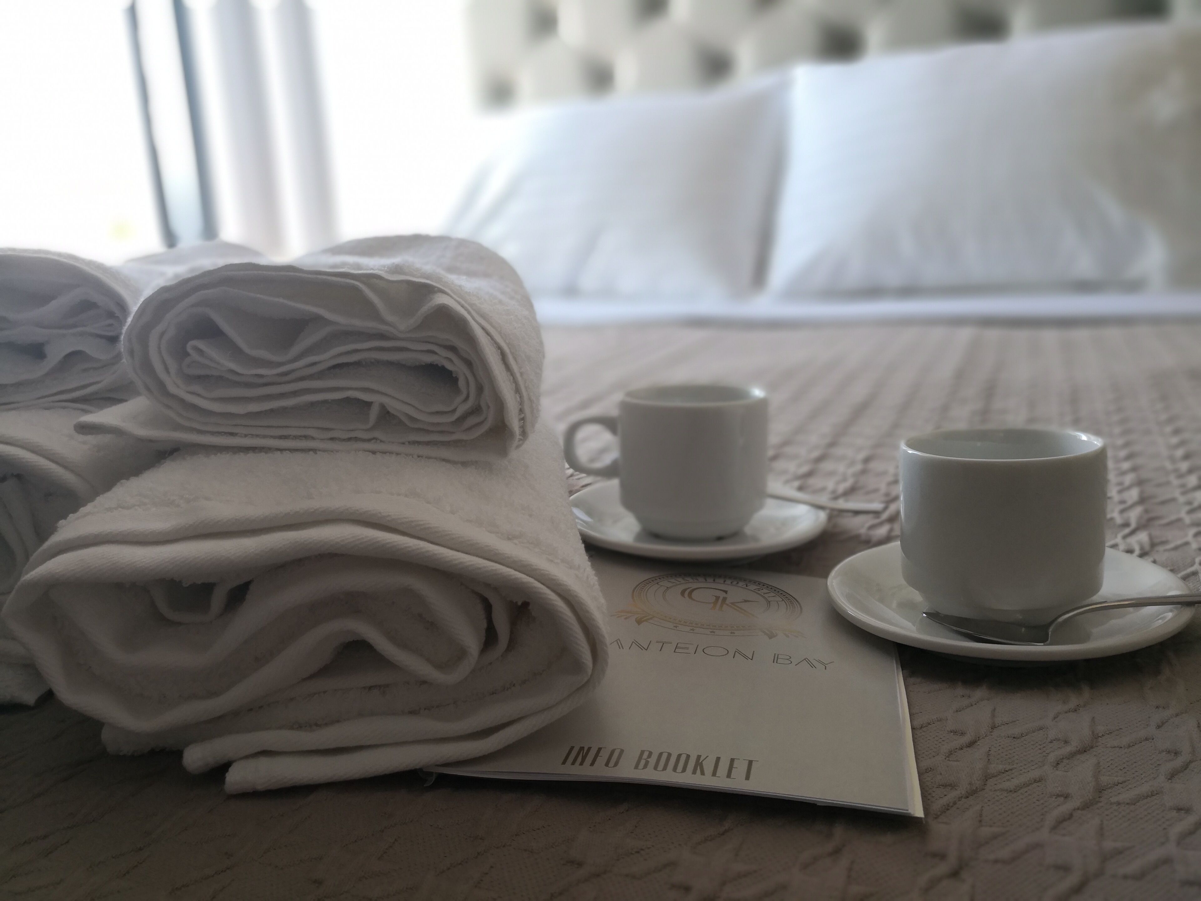 Una cama bien hecha con sábanas blancas, dos tazas blancas sobre platillos y un folleto titulado 'INTEION BAY INTO BOOKLET' sobre una colcha con textura.