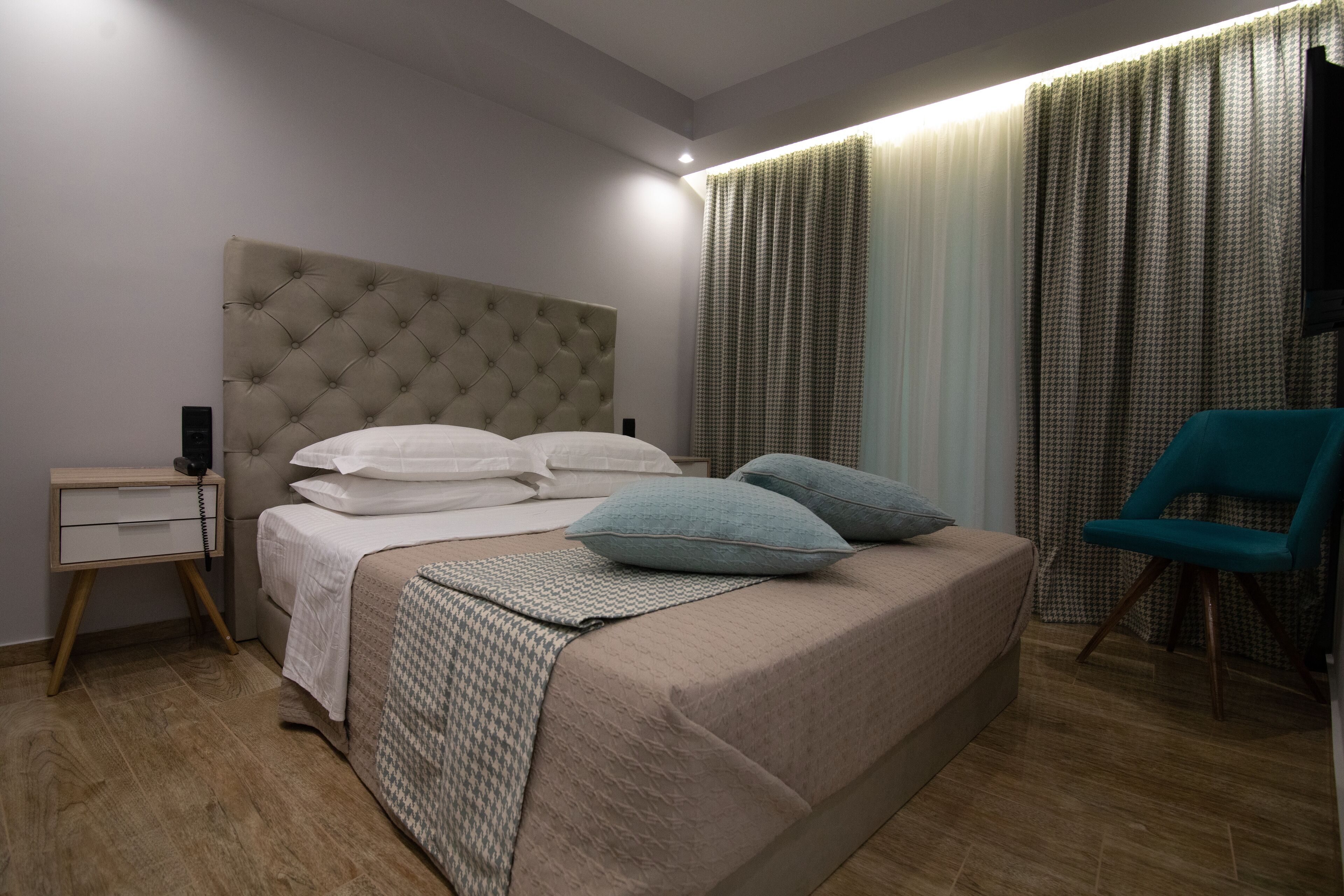 A bedroom with a tufted headboard, a bed with white and patterned linens, a teal chair, and a nightstand with a lamp.